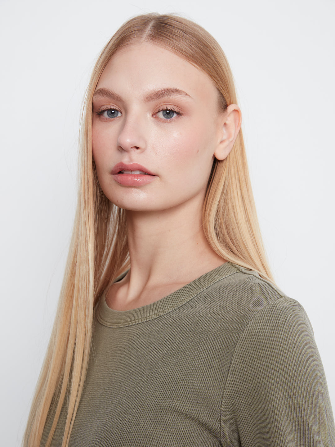 A close-up portrait of a blonde woman wearing a ribbed crewneck top in olive green, set against a plain light gray background.