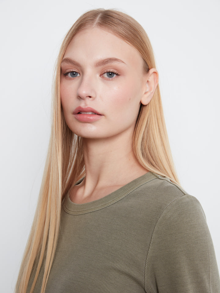 A close-up portrait of a blonde woman wearing a ribbed crewneck top in olive green, set against a plain light gray background.