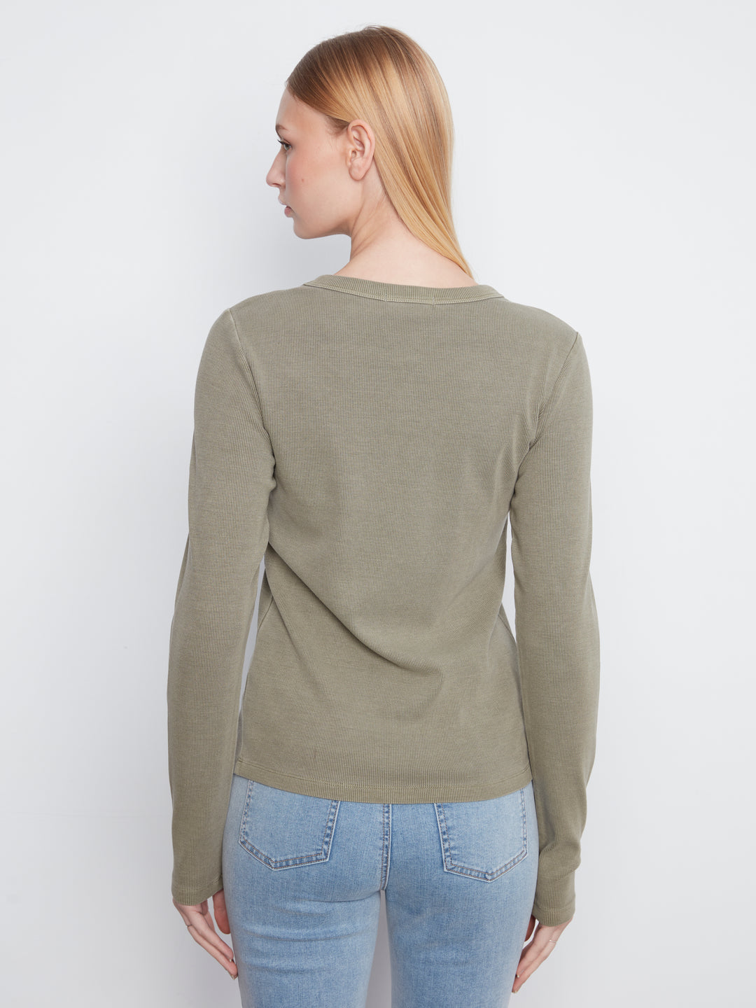A woman with blonde hair is shown from the back wearing a ribbed long-sleeve knit sweater in a muted olive green color, paired with light blue jeans, against a plain light gray background.
