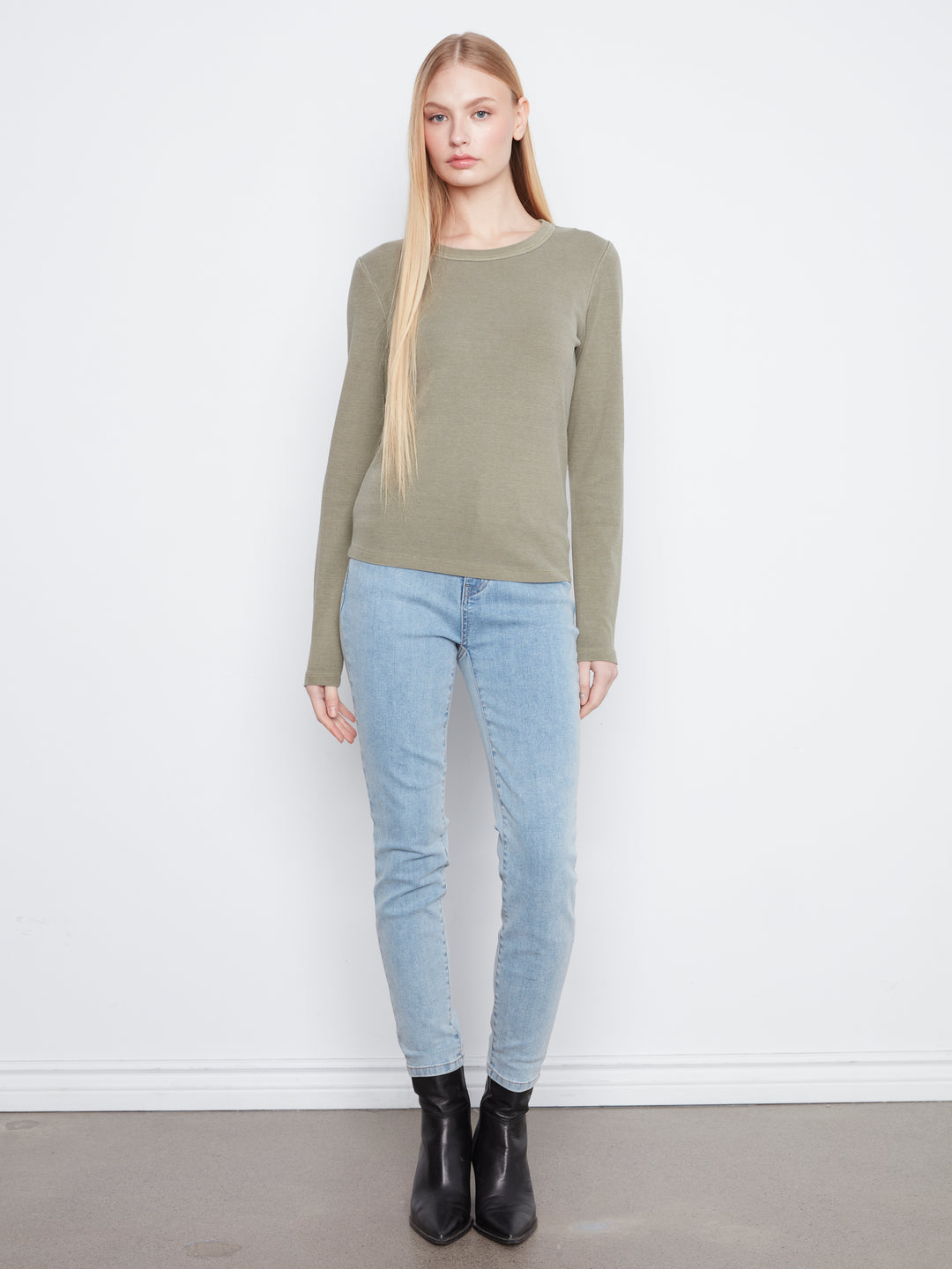 A woman stands against a white wall wearing a long-sleeve crewneck sweater in olive green, light-wash blue skinny jeans, and black ankle boots.