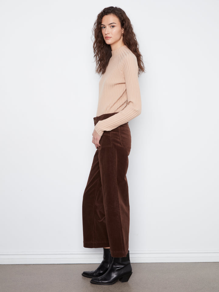 Woman wearing a beige sweater and brown velvet pants against a white wall.