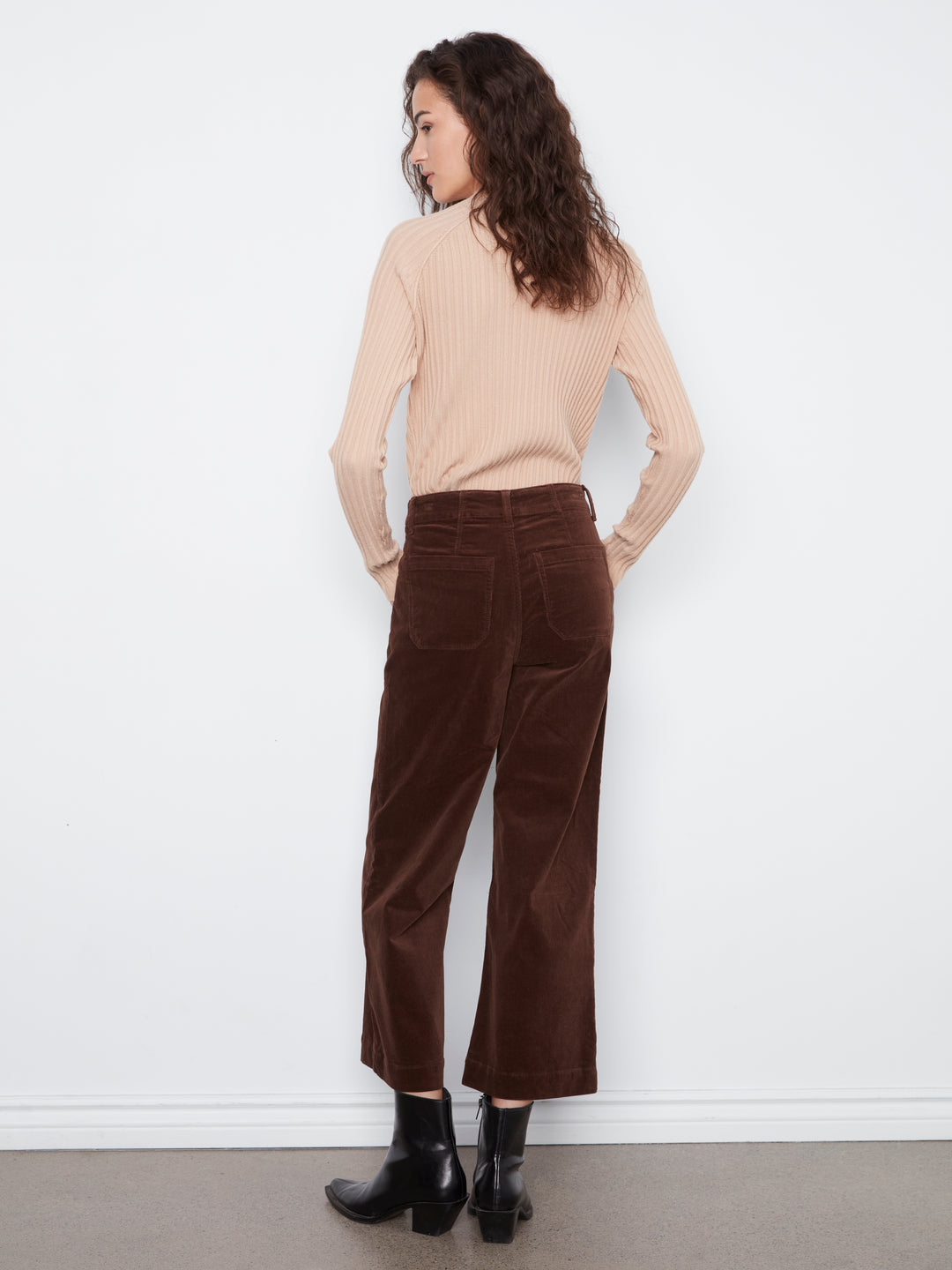Woman wearing a beige sweater and brown velvet pants against a white wall.