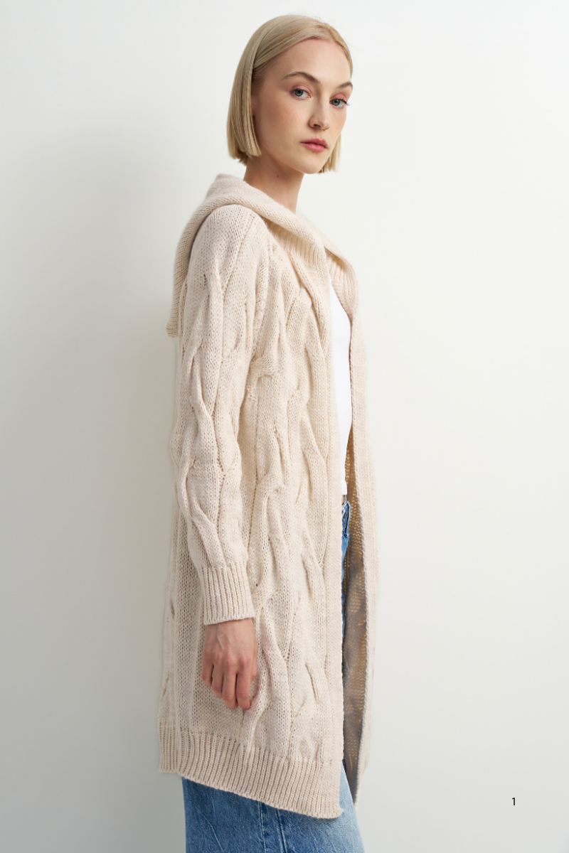 A woman wearing a long cream cable-knit hooded cardigan over a white top and blue jeans, standing against a plain off-white studio background.