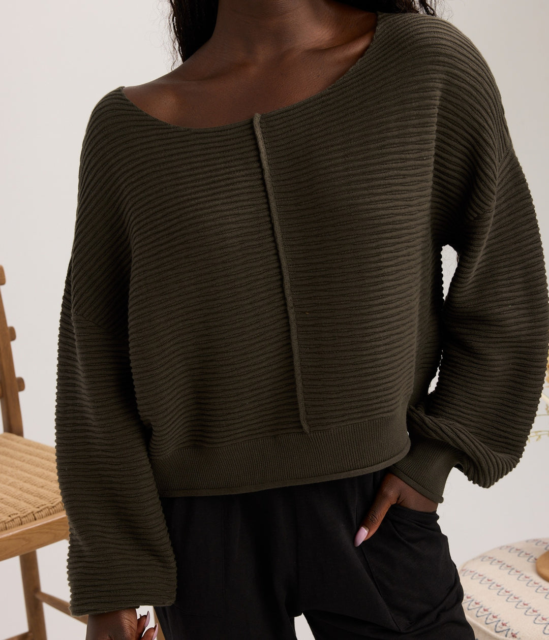 A person wearing a ribbed knit long-sleeve sweater in dark olive green, paired with black pants, shown in a styled indoor setting.