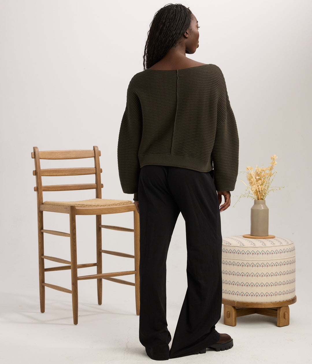A model is shown from behind wearing an olive green wide-neck ribbed sweater and black wide-leg trousers, standing in a studio setting next to a wooden chair and a patterned ottoman.