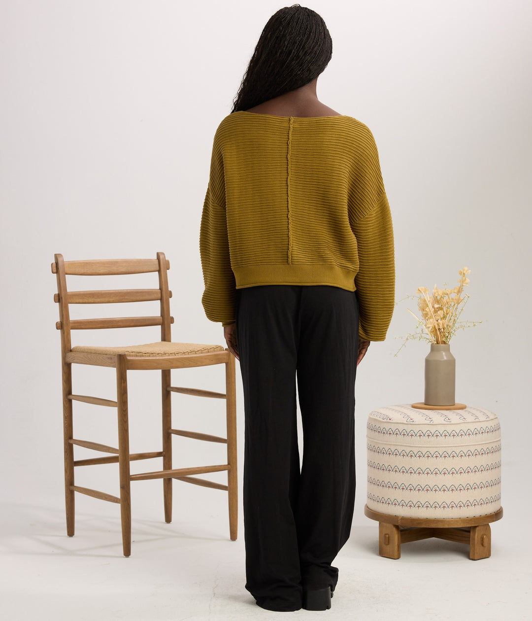 A woman stands with her back to the camera wearing a ribbed mustard yellow sweater and black pants, posed in a studio with a wooden chair, a patterned ottoman, and a small vase of dried flowers.