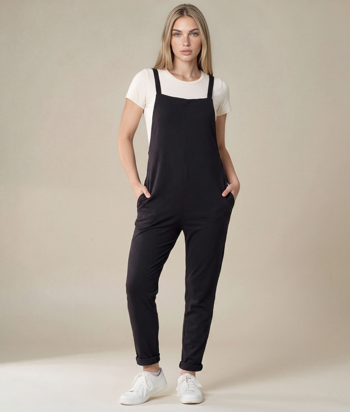 A woman modeling black overall-style pants with a white short-sleeve top and white sneakers, standing against a plain beige studio background.