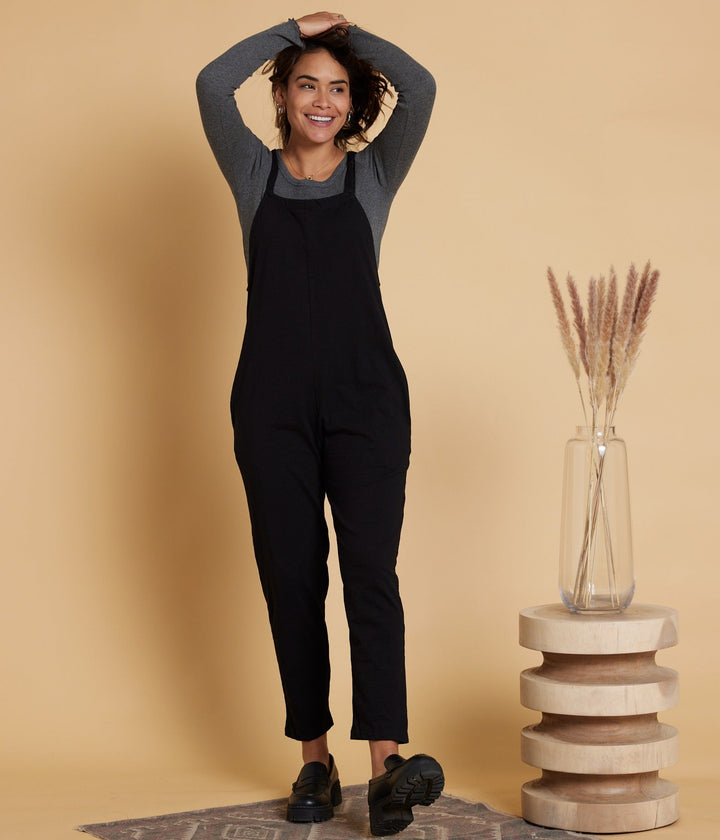 Woman in black overalls standing next to a wooden stool with a vase on it against a beige wall.