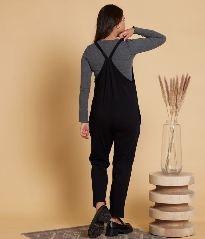 Woman wearing black overalls against a beige wall with a wooden stool and vase.
