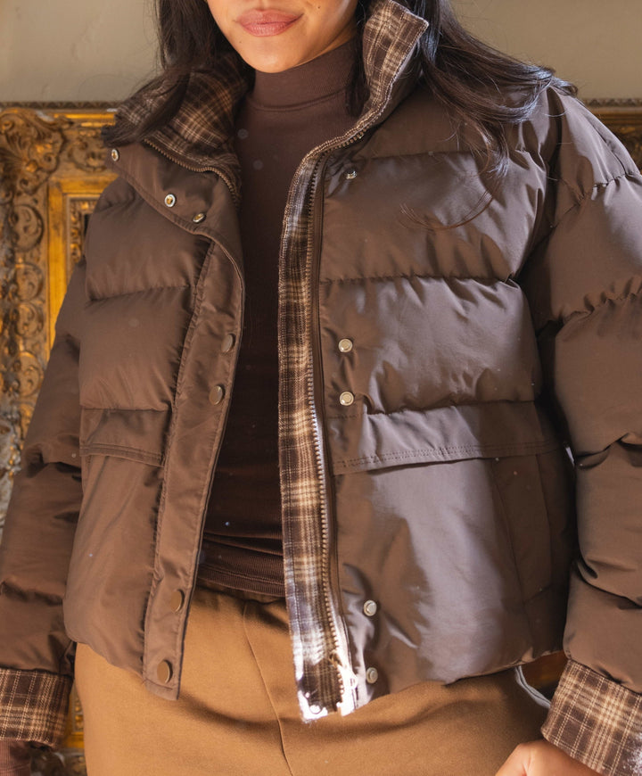 A woman wearing a cropped brown puffer jacket with a plaid collar, cuffs, and front plaid lining, paired with a dark brown turtleneck and tan pants, posed in front of an ornate gold-framed mirror.