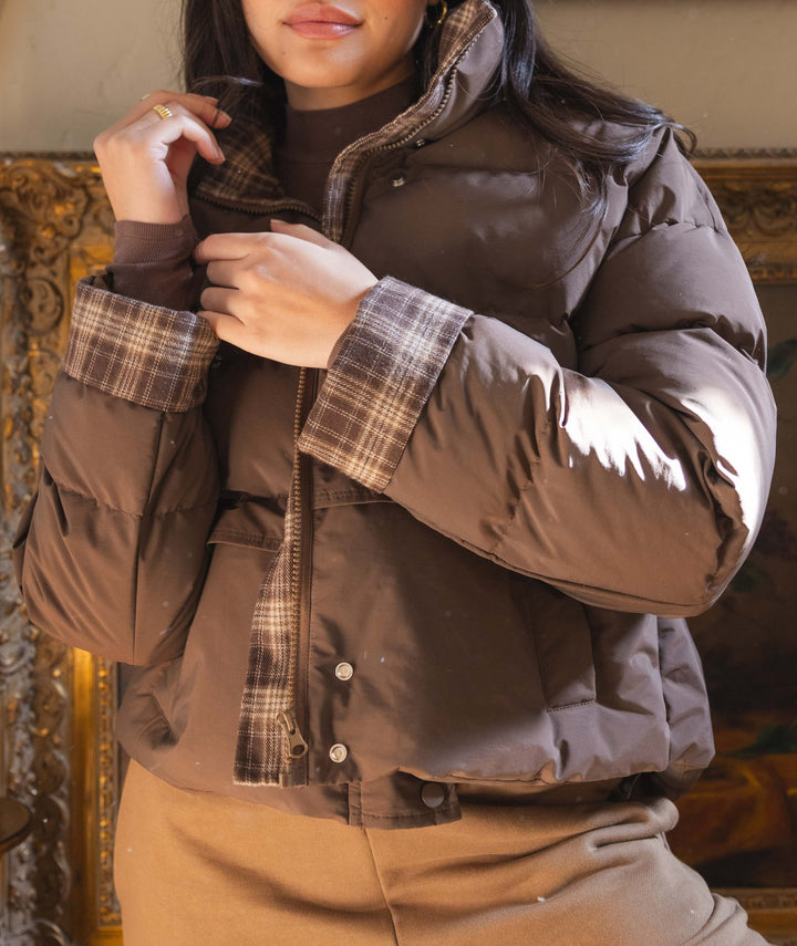 A person wearing a brown puffer jacket with plaid trim and a brown top, standing indoors in front of a large ornate gold-framed mirror.