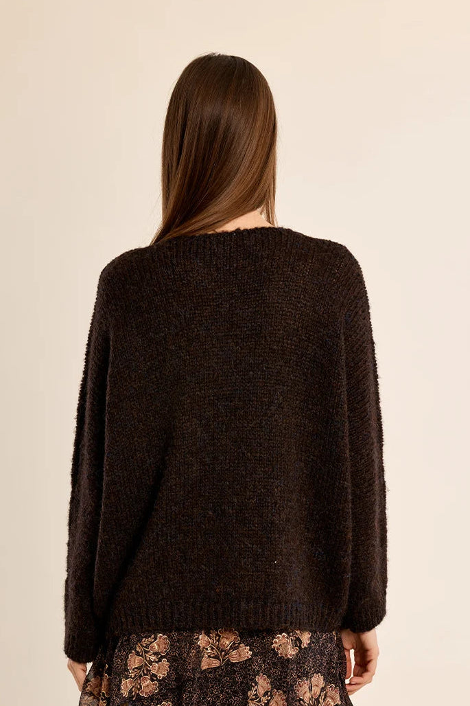 A woman shown from the back wearing a dark brown fuzzy knit sweater and a dark floral skirt, set against a plain beige studio background.