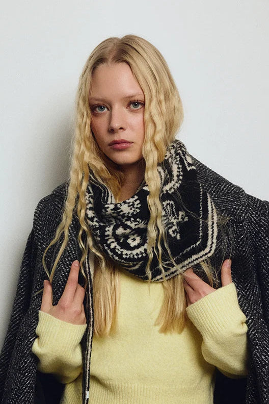 A blonde model wearing a pale yellow knit sweater, a black and white patterned bandana scarf, and a dark gray textured overcoat, posing against a light gray background.