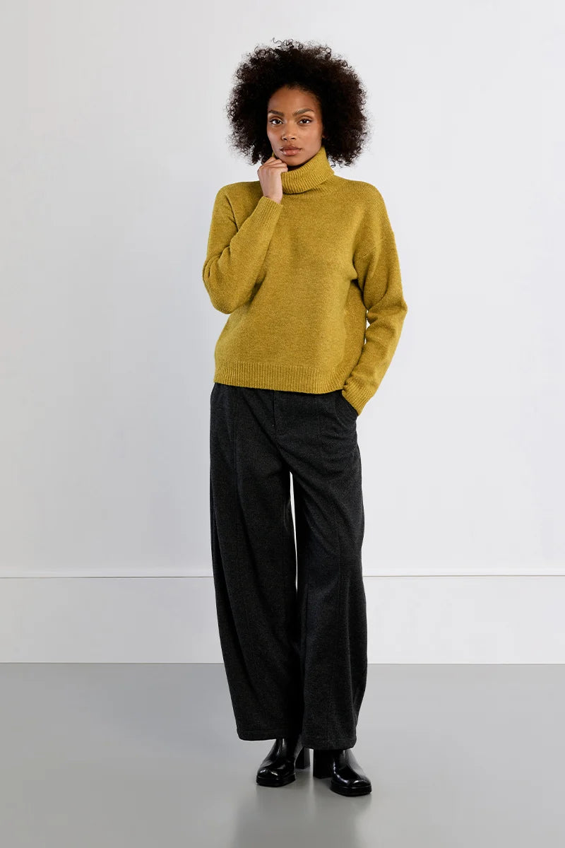 A model with curly dark hair stands in a studio wearing a mustard yellow turtleneck sweater, black wide-leg trousers, and black leather shoes against a light gray background.