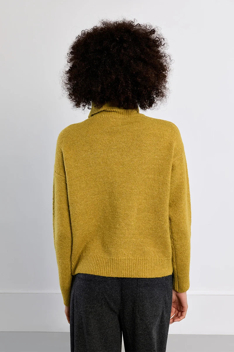 A person viewed from the back wearing a mustard yellow knit sweater and dark gray trousers against a plain light gray wall.