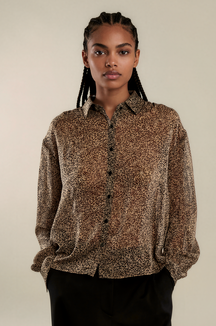 A woman modeling a brown and beige leopard-print button-up shirt with black pants, standing against a plain beige studio background.