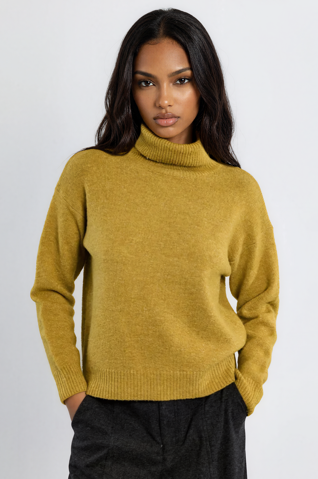 A woman wearing a mustard yellow knit turtleneck sweater and dark charcoal trousers, standing against a plain light gray studio background.