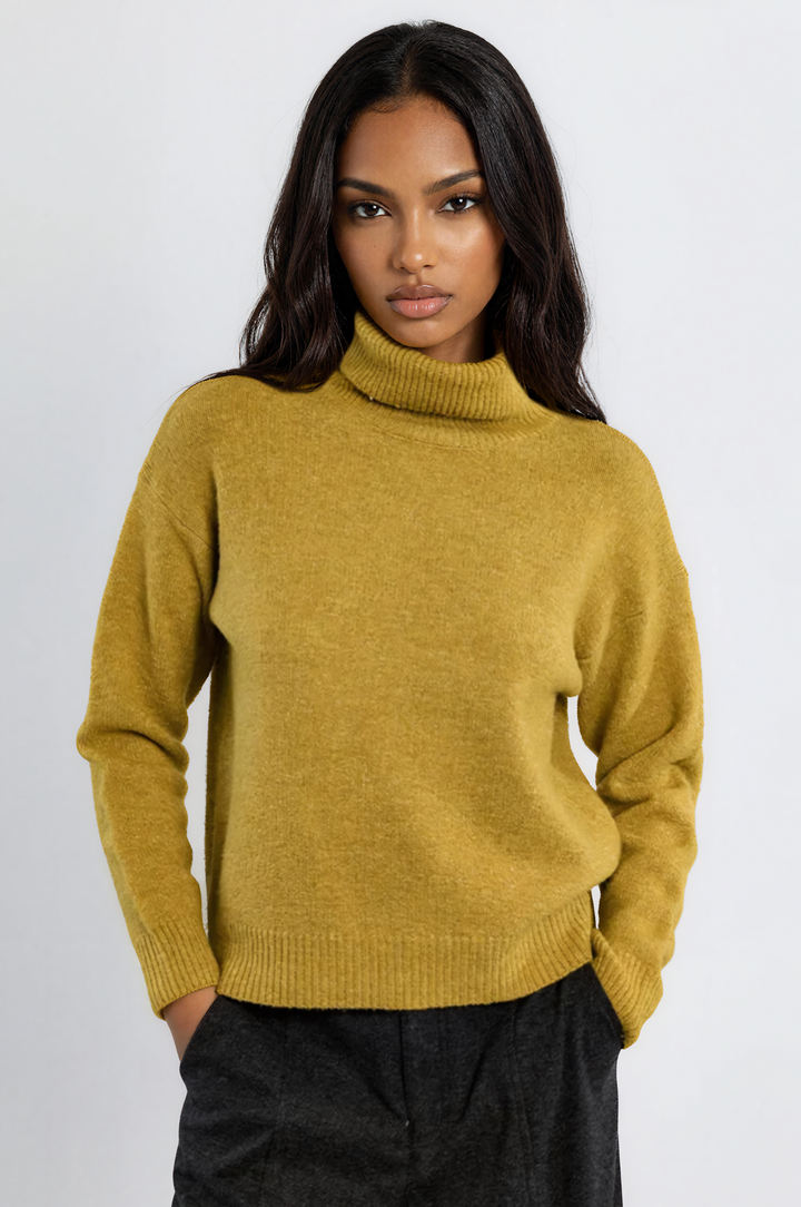 A woman wearing a mustard yellow knit turtleneck sweater and dark charcoal trousers, standing against a plain light gray studio background.