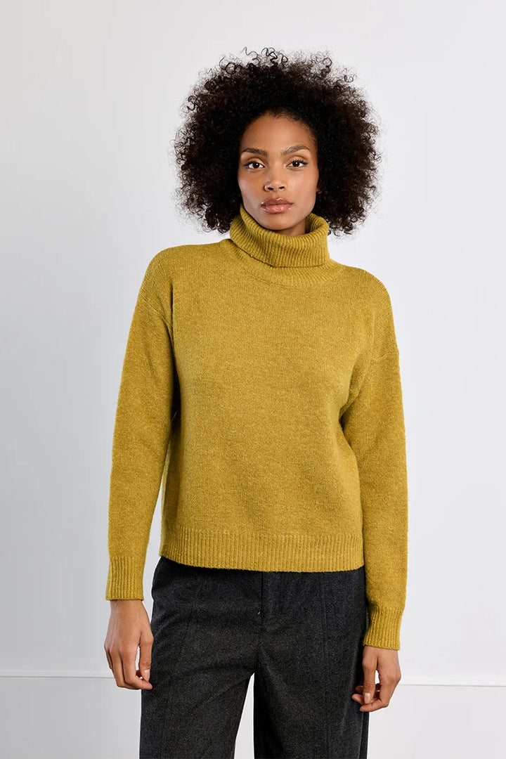 A woman with curly dark hair wearing a mustard yellow ribbed turtleneck sweater and dark charcoal trousers, standing against a plain light gray studio background.