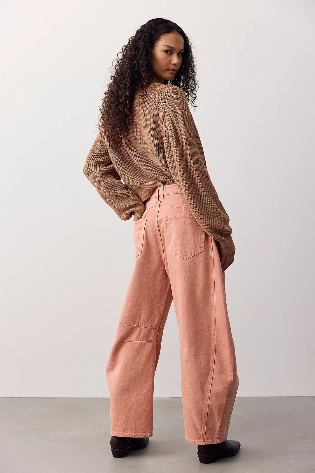 A woman stands in a studio wearing a brown ribbed knit sweater and wide-leg dusty pink pants, photographed from the back with her head turned over her shoulder.