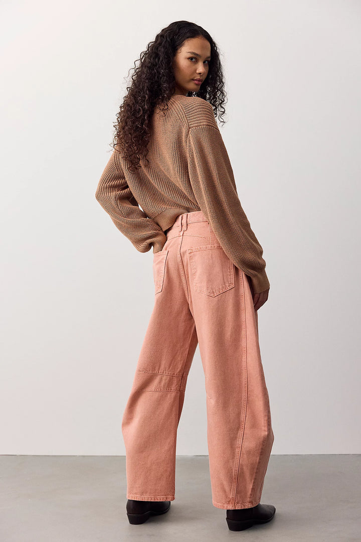 A woman stands in a studio wearing a brown ribbed knit sweater and wide-leg dusty pink pants, photographed from the back with her head turned over her shoulder.