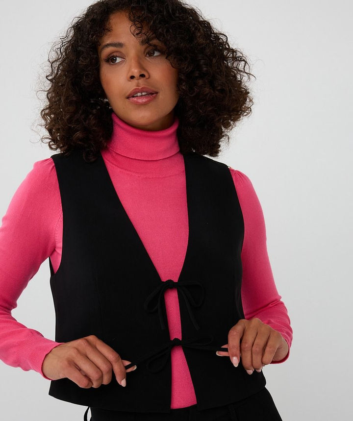 Woman wearing a black vest over a pink turtleneck against a white background