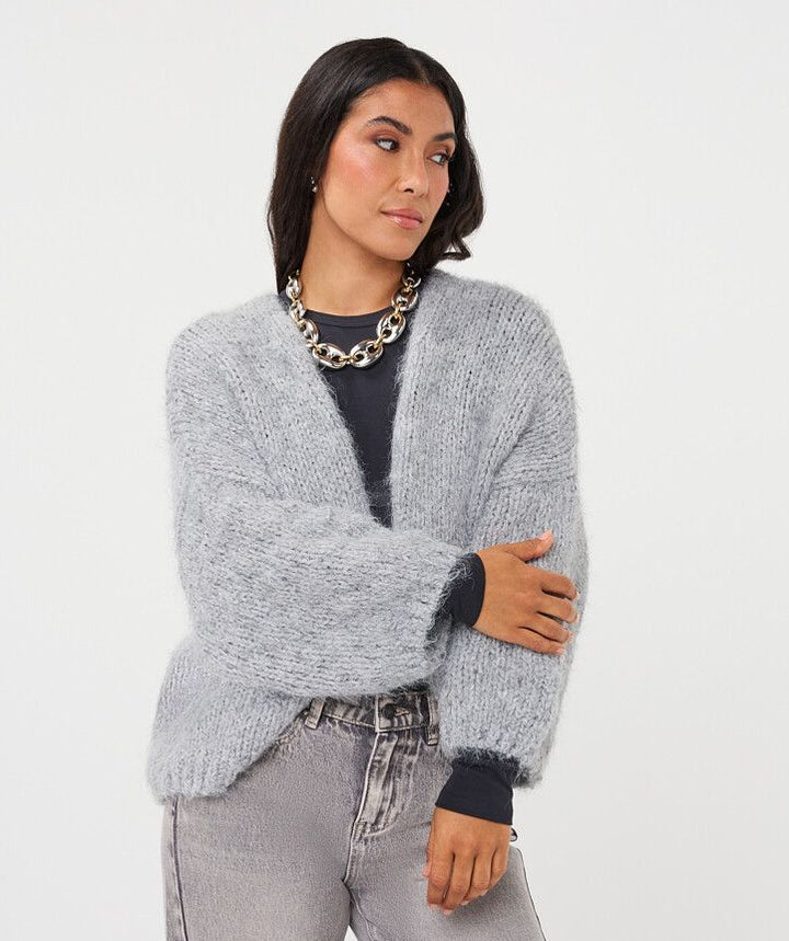 A woman modeling a light gray fuzzy open-front cardigan sweater over a black top and gray jeans, standing against a plain light gray studio background.