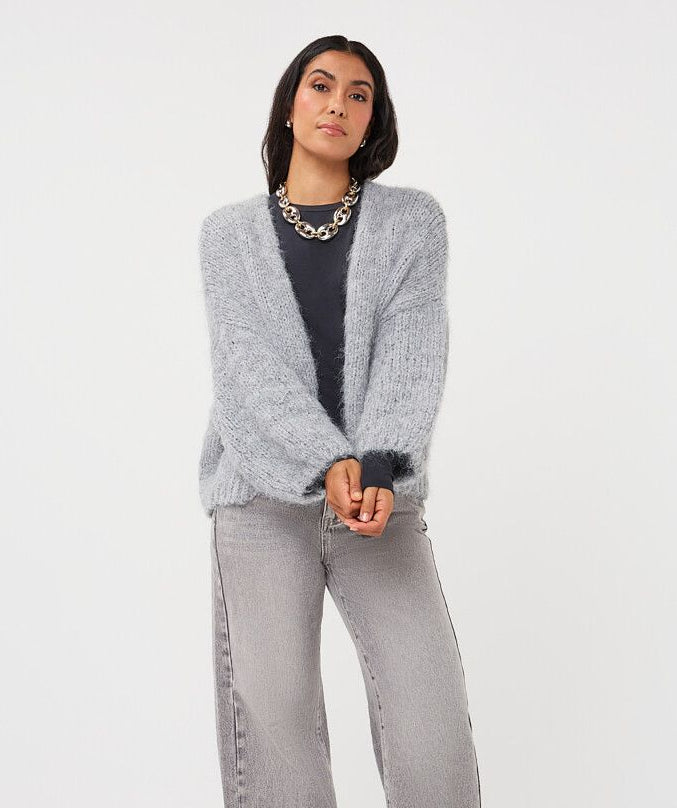 A woman modeling a light gray knit cardigan over a black top, paired with wide-leg gray jeans and black pointed shoes, against a plain white studio background.