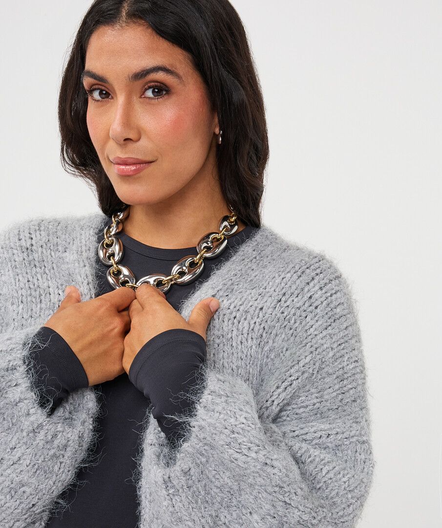 A woman wearing a chunky silver chain necklace over a black top and a fuzzy light gray cardigan, posing against a plain light gray background.