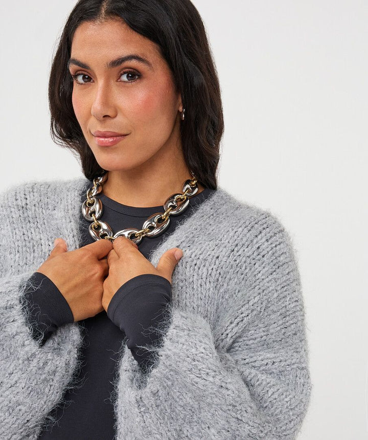 A woman wearing a chunky silver chain necklace over a black top and a fuzzy light gray cardigan, posing against a plain light gray background.