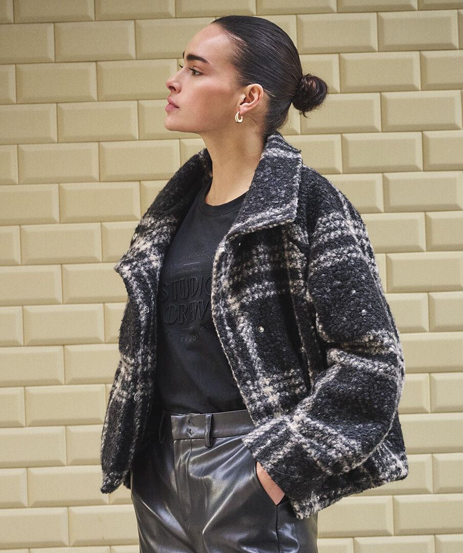 A woman wearing a black and white plaid bouclé jacket over a black t-shirt and black leather pants, standing against a beige brick wall.