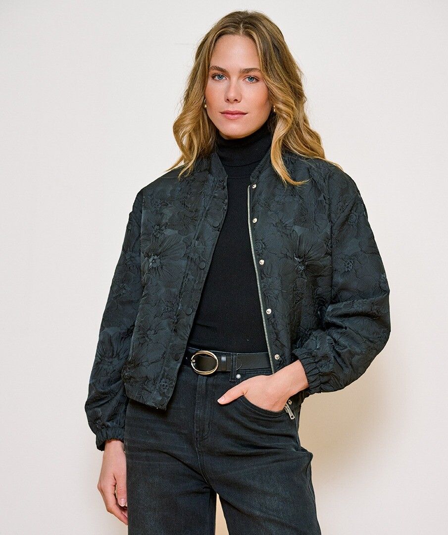 A woman modeling a dark textured bomber jacket with a floral jacquard pattern, worn over a black turtleneck and dark jeans against a plain light background.