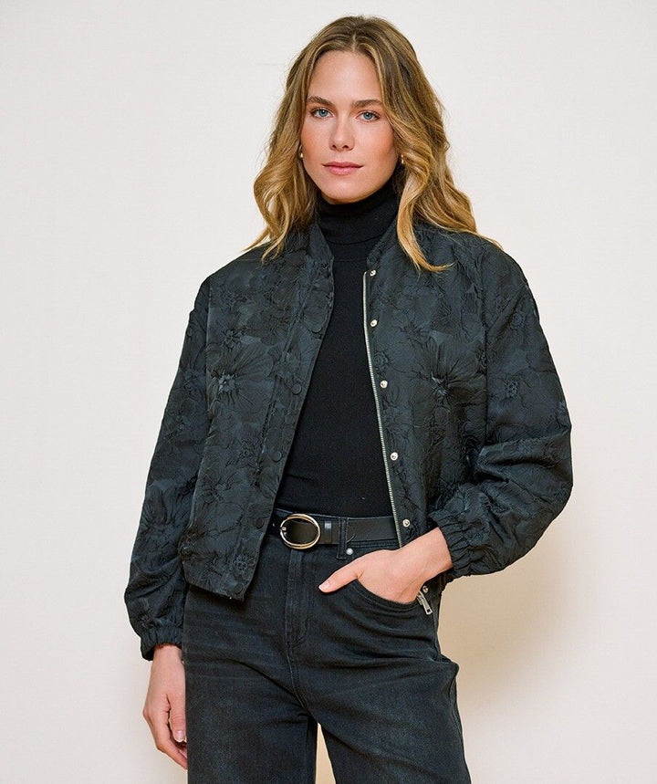 A woman modeling a dark textured bomber jacket with a floral jacquard pattern, worn over a black turtleneck and dark jeans against a plain light background.