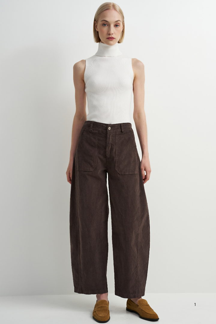 A model stands against a plain light gray studio background wearing a white sleeveless mock-neck top, dark brown wide-leg corduroy pants, and brown suede loafers.