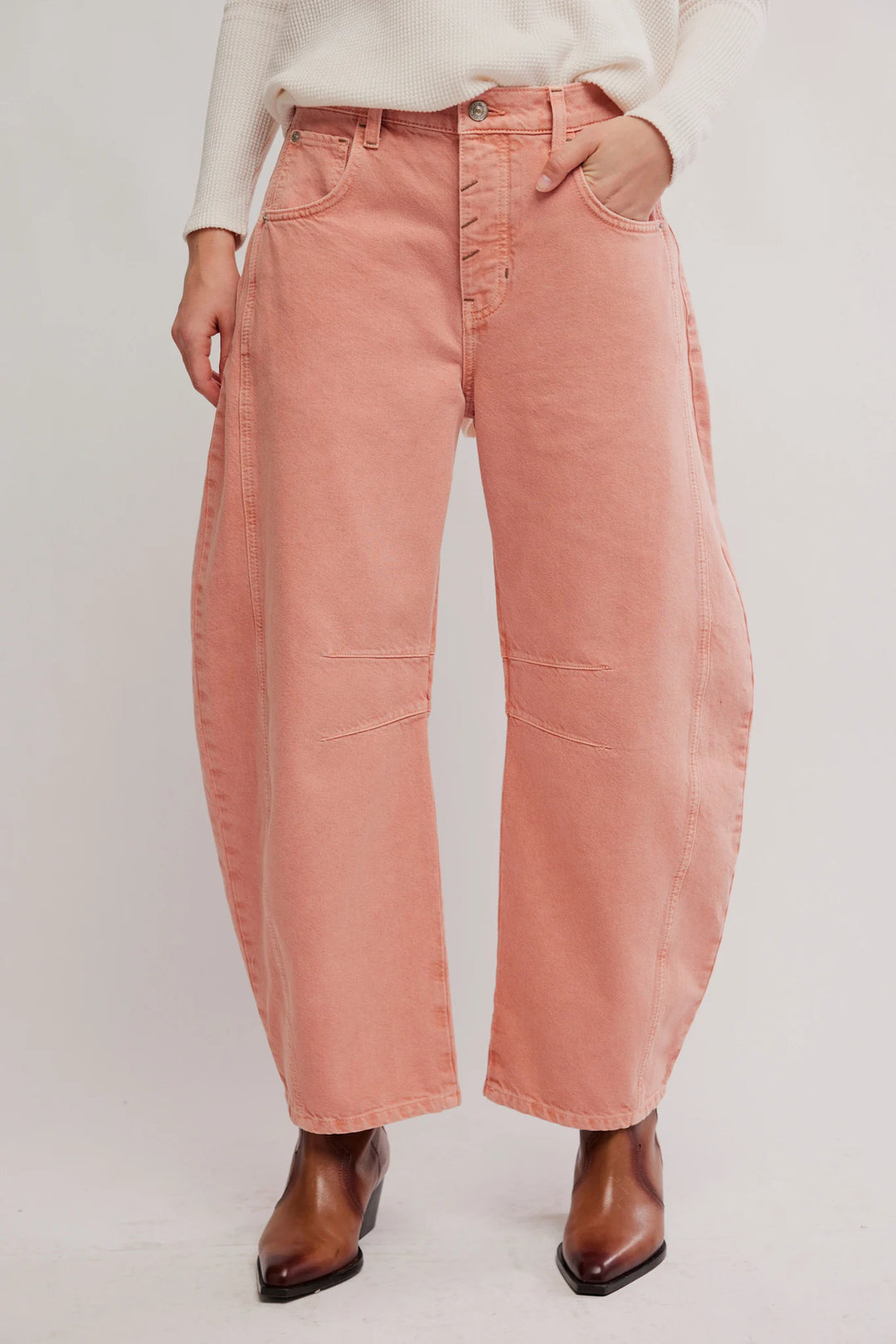 A person wearing oversized dusty pink wide-leg jeans with a button fly, paired with a cream top and brown pointed-toe boots, photographed against a plain light background.