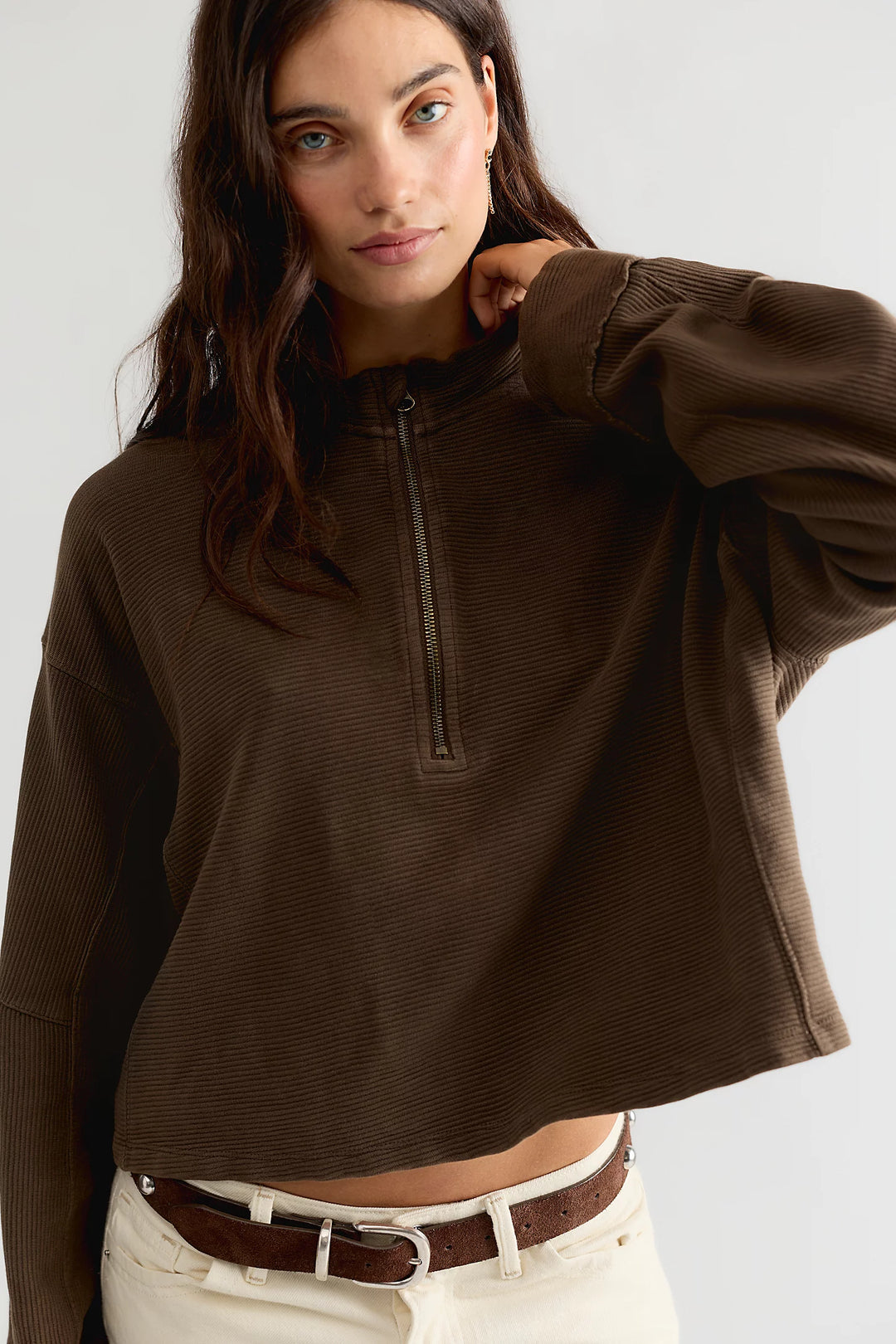 A woman wearing a dark brown ribbed long-sleeve half-zip pullover top with cream pants and a brown belt, photographed against a light gray studio background.