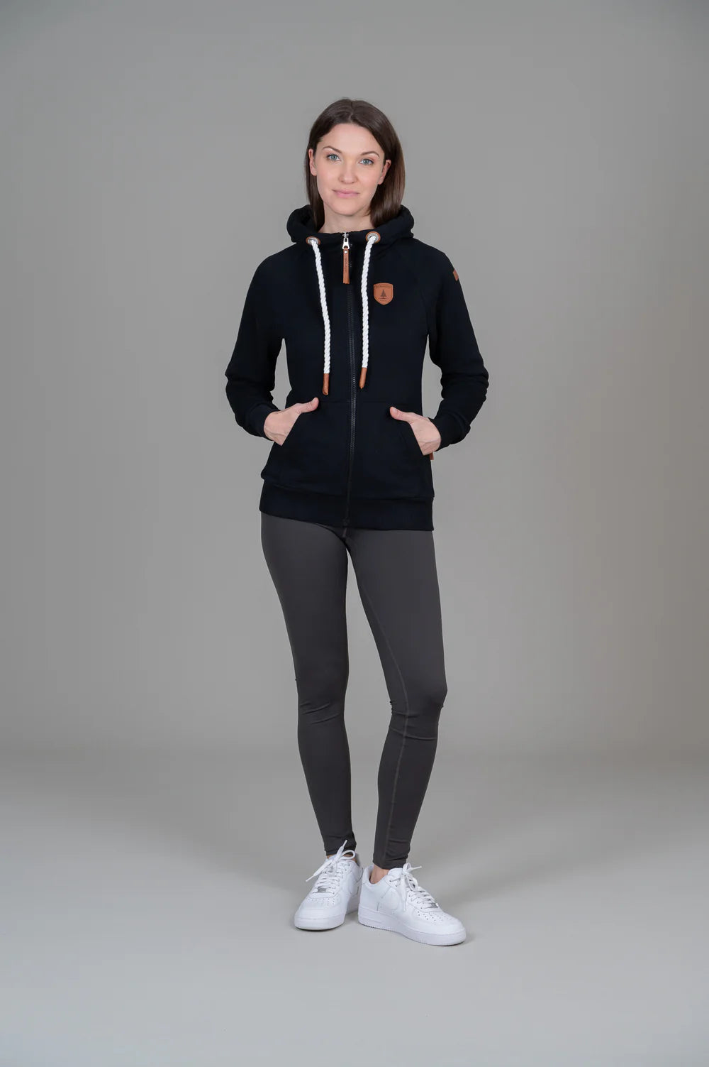Person wearing a black hoodie with a logo, gray leggings, and white sneakers on a plain background