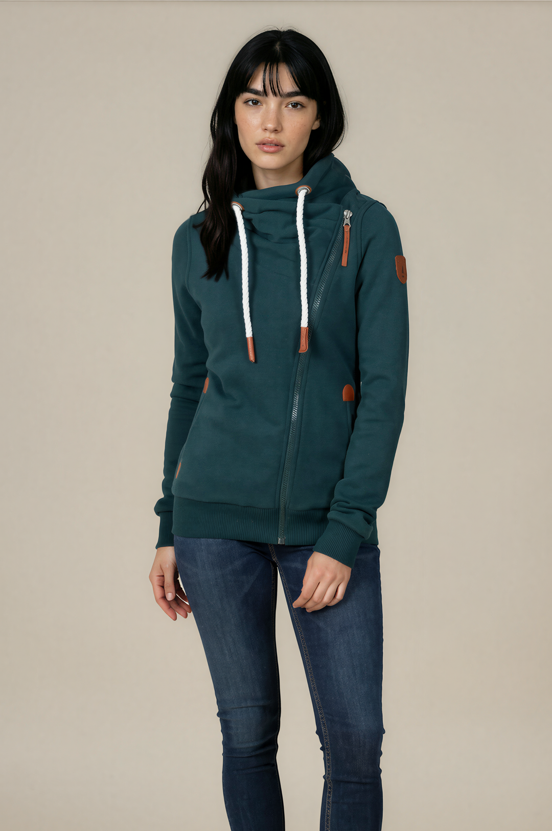 A woman modeling a dark teal green zip-up hoodie with white drawstrings and brown leather-like accents, paired with dark blue skinny jeans and white sneakers, against a solid beige studio background.