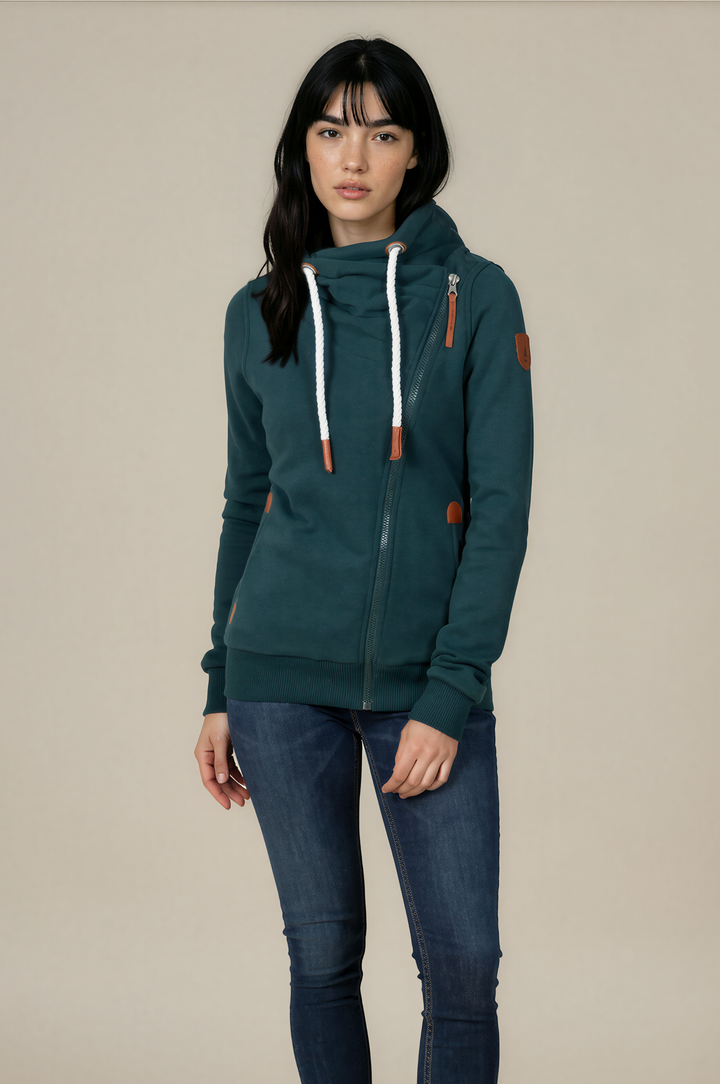 A woman modeling a dark teal green zip-up hoodie with white drawstrings and brown leather-like accents, paired with dark blue skinny jeans and white sneakers, against a solid beige studio background.
