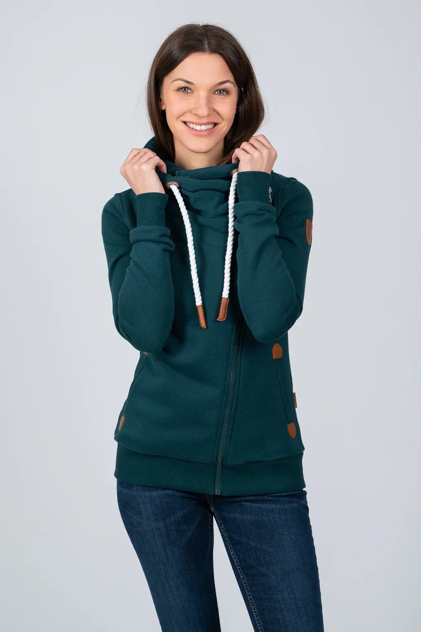 A woman wearing a dark teal zip-up hoodie with white braided drawstrings and brown accents, paired with dark blue jeans, against a light gray studio background.