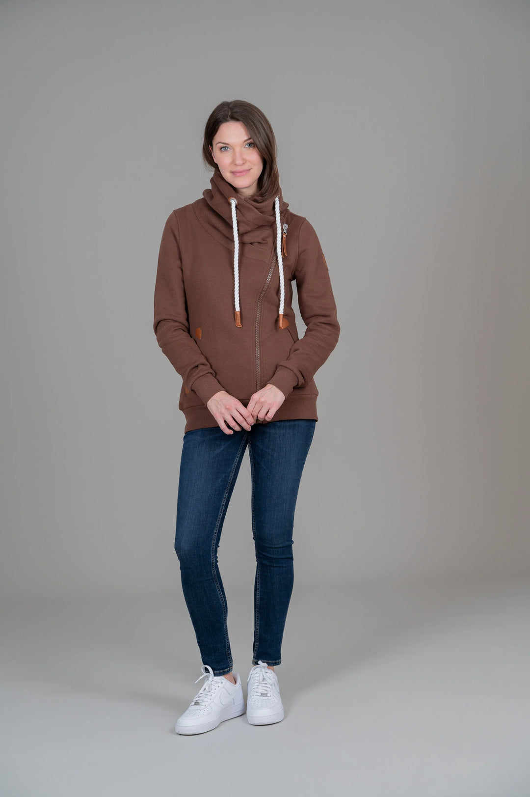 A woman modeling a brown zip-up hoodie with white drawstrings, paired with dark blue skinny jeans and white sneakers, standing against a neutral gray studio background.