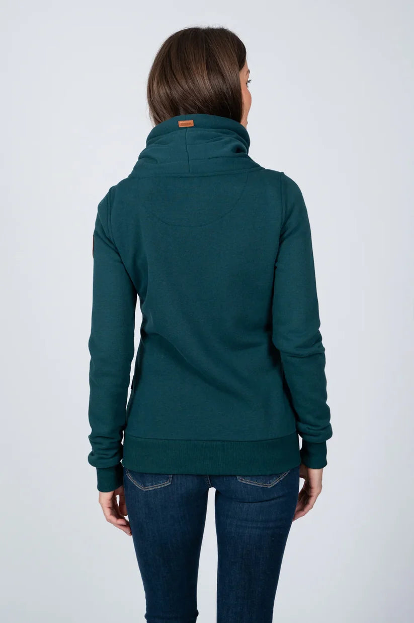 A woman viewed from the back wearing a dark teal green mock neck pullover hoodie with dark blue jeans, set against a plain light gray studio background.