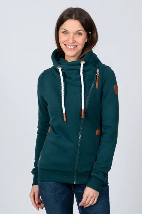 A woman modeling a dark teal green zip-up hoodie with white drawstrings and brown accents, paired with dark blue skinny jeans and white sneakers, against a light gray studio background.