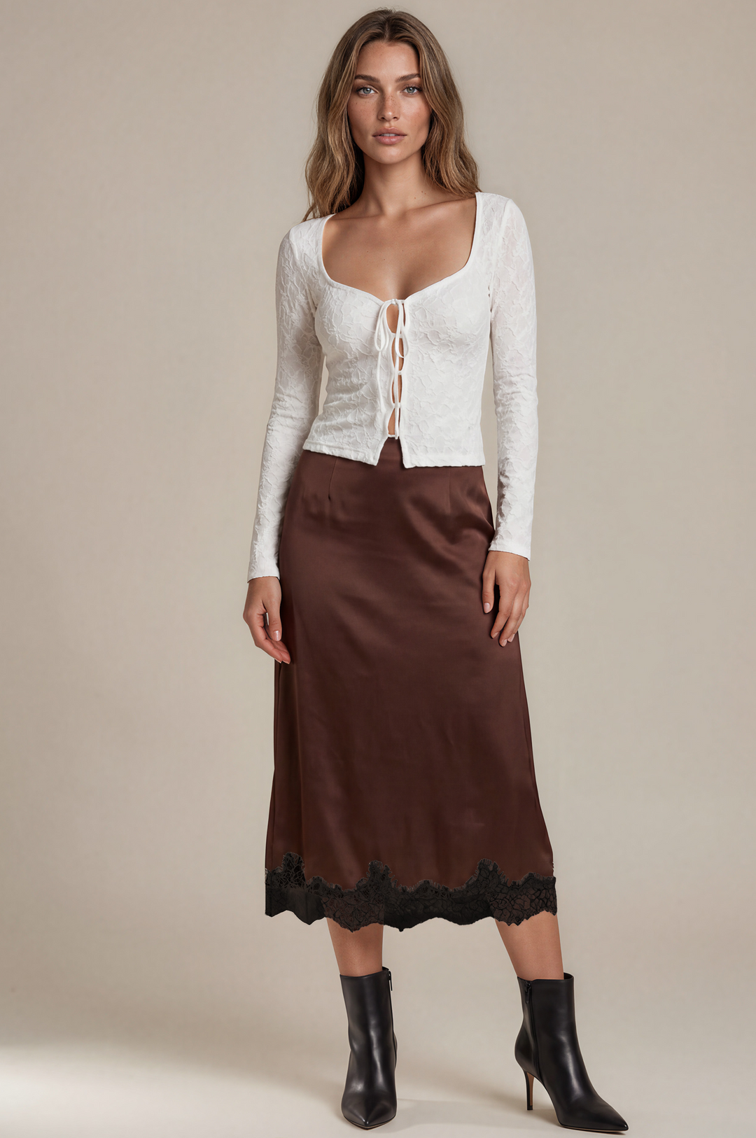 A woman modeling a white lace long-sleeve crop top with lace-up front and a brown midi skirt with black lace hem, paired with black ankle boots, against a beige studio background.