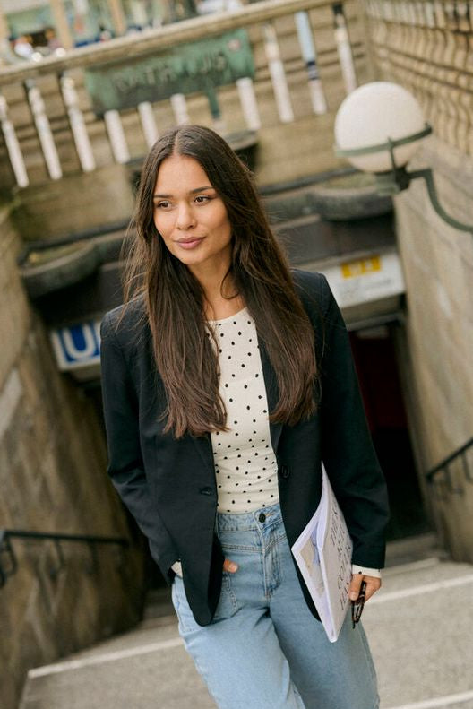A woman walking down outdoor stone stairs wearing a black blazer, a white top with black polka dots, light blue denim shorts, and black boots, holding a white folder.