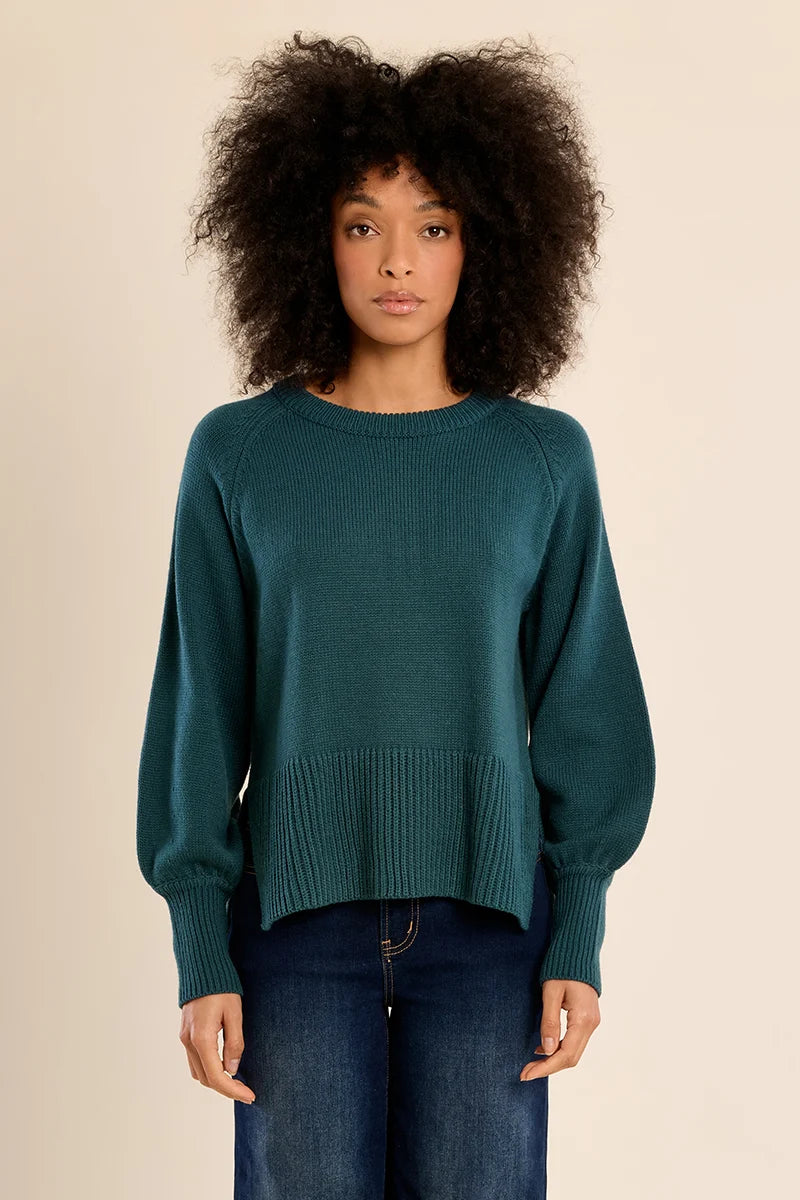 A woman with voluminous dark curly hair wearing a teal green long-sleeve knit sweater and dark blue jeans, standing against a solid beige studio background.