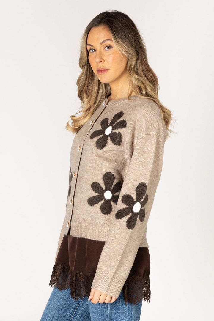 A woman modeling a beige knit cardigan with large dark brown flower appliqués and a dark brown lace-trimmed hem, paired with blue jeans against a plain light background.