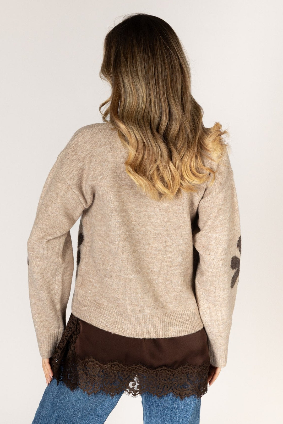 A woman viewed from behind wearing a beige knit sweater with dark brown lace trim at the hem and dark brown paw-print details on the sleeves, paired with blue jeans against a plain light background.