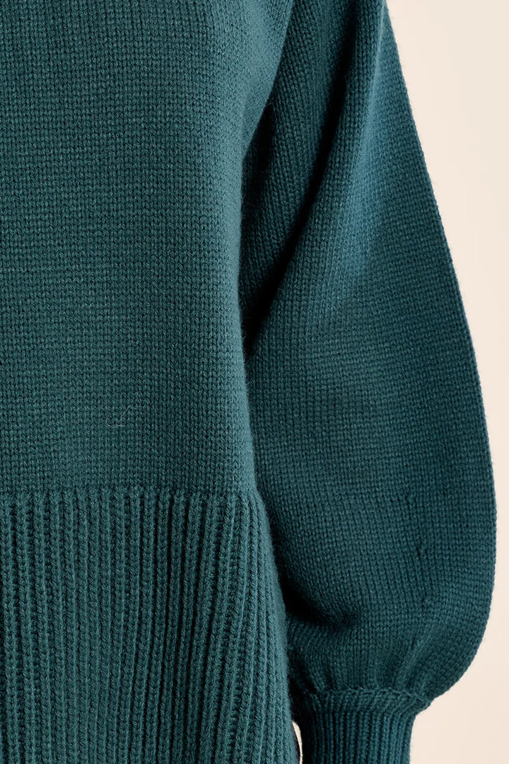 Close-up of a dark teal knit sweater showing the ribbed sleeve cuff and textured fabric against a light beige background.