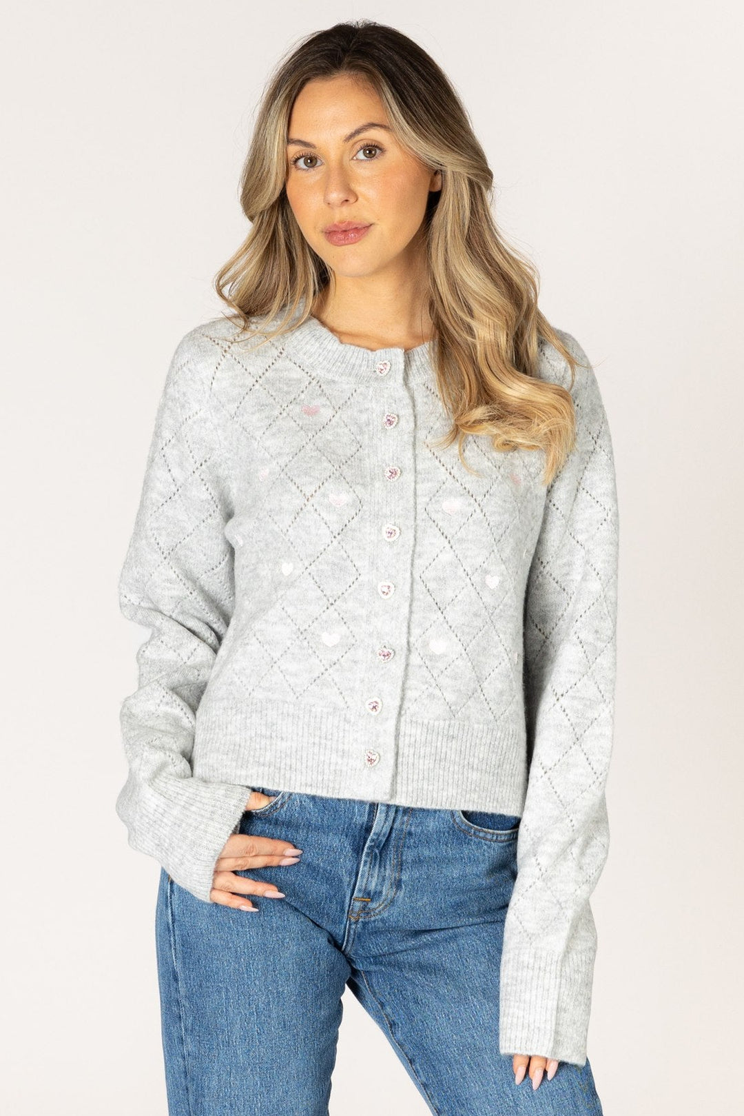 A woman modeling a light gray textured knit cardigan sweater with a diamond pattern and button closure, paired with blue jeans, against a plain light gray studio background.